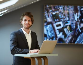 Portrait of handsome bearded businessman looking at camera and smiling while posing at standing table in futuristic office interior with city map in background, copy space